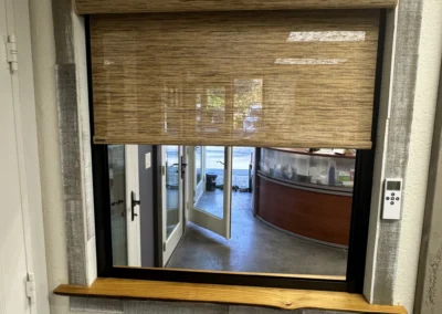 A partially open woven shade covers a window with a wooden ledge, revealing a view of a bright, modern interior space.