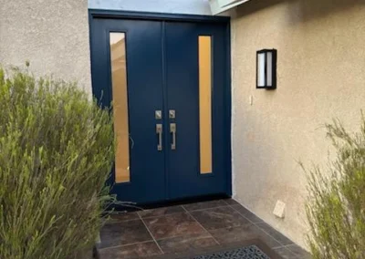 A modern navy blue double door with glass panels, framed by green shrubs, a welcome mat, and a stylish light fixture above.