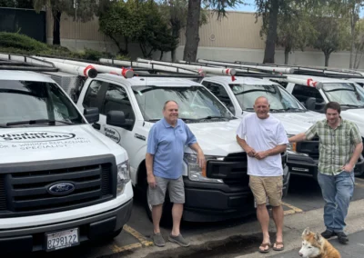 Four men stand beside parked work trucks, with tools on top, while a dog sits on the pavement nearby.